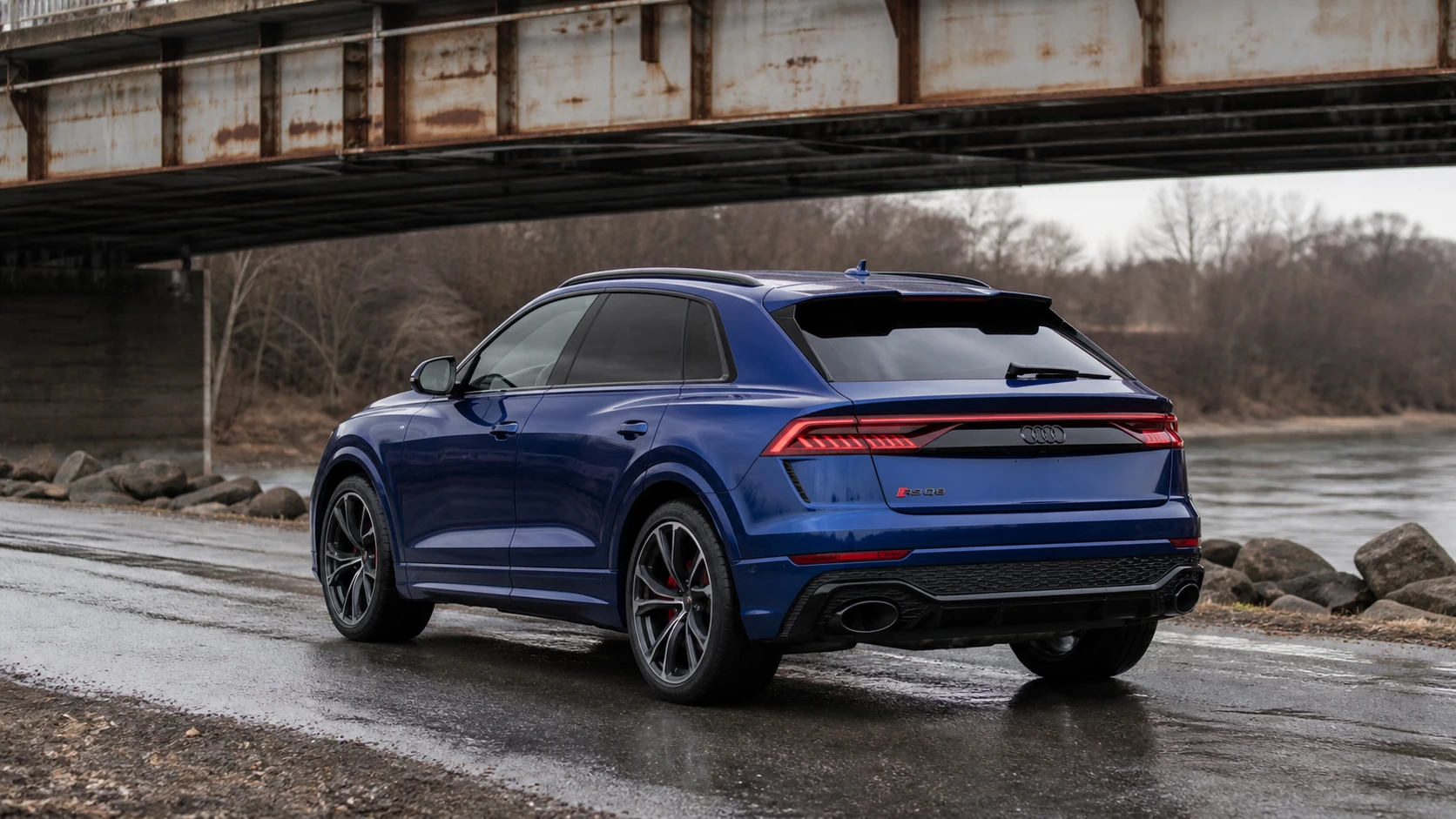 Blue Audi RS Q8 in a rear three-quarter view on a wet riverside road