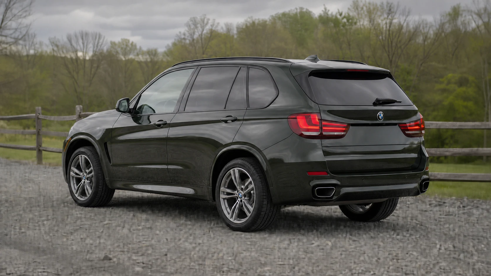 Dark olive F15 BMW X5 in a rear three-quarter view on a wooded gravel road