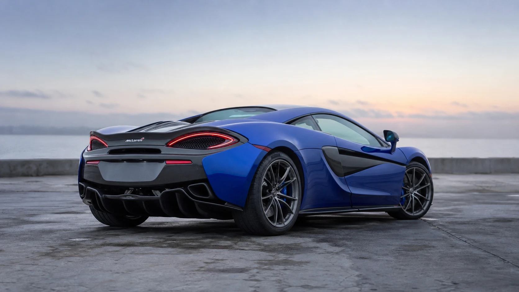 Blue McLaren 570S in a rear three-quarter view on damp harbor-edge concrete